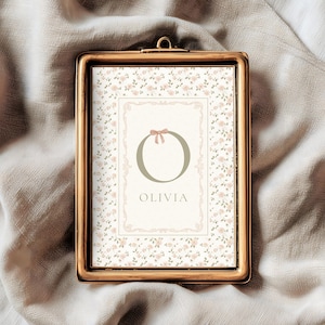 Pu&ograve; includere: Una stampa incorniciata con una cornice color oro. La stampa presenta un motivo floreale e un disegno centrale con la lettera "O", un fiocco e il nome "Olivia". Lo sfondo &egrave; di un colore morbido e neutro.