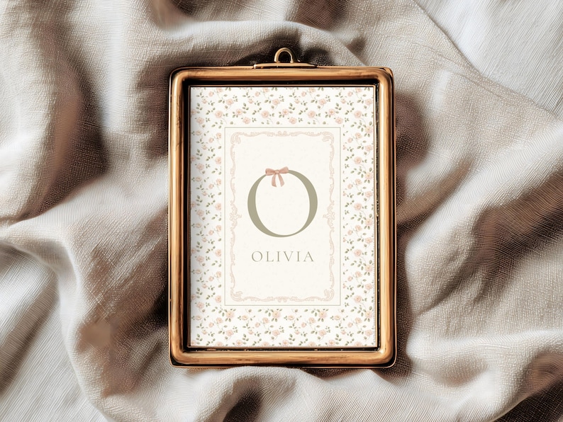 Pu&ograve; includere: Una stampa incorniciata con una cornice color oro. La stampa presenta un motivo floreale e un disegno centrale con la lettera "O", un fiocco e il nome "Olivia". Lo sfondo &egrave; di un colore morbido e neutro.