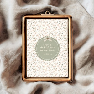 Op de afbeelding: Een rechthoekige, goudkleurige ingelijste print met een bloemenpatroon achtergrond. De print heeft een cirkelvormig ontwerp met de tekst "Trust in the Lord with all your heart." Het frame heeft een kleine lus aan de bovenkant om op te hangen.