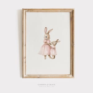 May include: A framed watercolour painting of two bunnies wearing pink dresses. The bunnies are standing side-by-side and looking at each other. The painting is set against a white background.