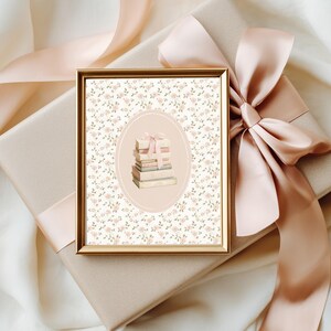 May include: A framed print with a gold border, featuring a stack of books tied with a pink ribbon, set against a floral background. The print rests on a wrapped gift box with a large pink satin bow. The background is a soft, off-white fabric.