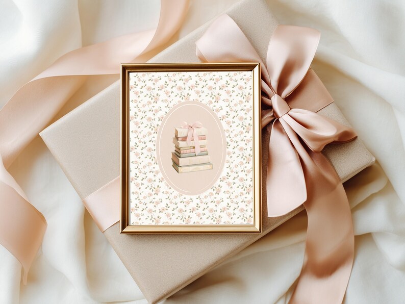May include: A framed print with a gold border, featuring a stack of books tied with a pink ribbon, set against a floral background. The print rests on a wrapped gift box with a large pink satin bow. The background is a soft, off-white fabric.