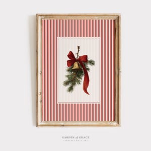 Op de afbeelding: Een ingelijste wandkunst met een vintage kerstthema. Het toont een gouden bel, een rode strik en dennentakken tegen een rood-wit gestreepte achtergrond. De lijst is van licht hout. De tekst "GARDEN OF GRACE" staat onderaan.