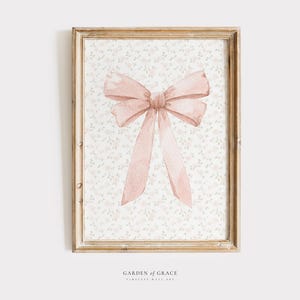 May include: A framed watercolour print showing a large, soft pink bow. The bow is centred on a background of small, light pink floral patterns. The frame is a light brown colour. The text "GARDEN OF GRACE TIMELESS WALL ART" is at the bottom.