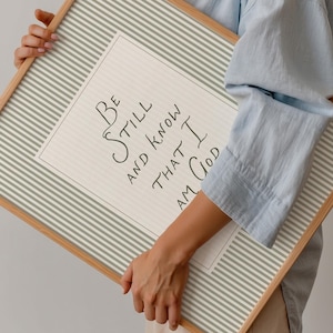 May include: A framed print with the text "Be Still and Know That I Am God." The print has a light beige background with a white and gray striped border. The frame is made of light-colored wood.