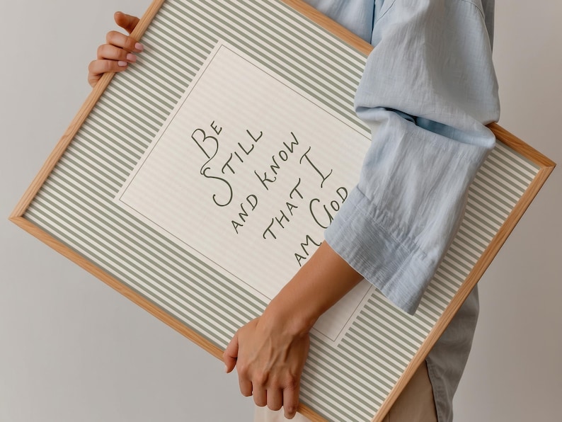 May include: A framed print with the text "Be Still and Know That I Am God." The print has a light beige background with a white and gray striped border. The frame is made of light-colored wood.