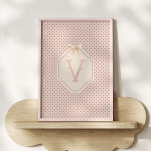 Puede incluir: Una impresi&oacute;n enmarcada con un fondo estampado rosa y blanco. La impresi&oacute;n tiene un marco rosa y un dise&ntilde;o central con la letra "V" en una fuente decorativa. Un peque&ntilde;o lazo adorna la parte superior del dise&ntilde;o. La impresi&oacute;n se muestra en una estanter&iacute;a de madera.