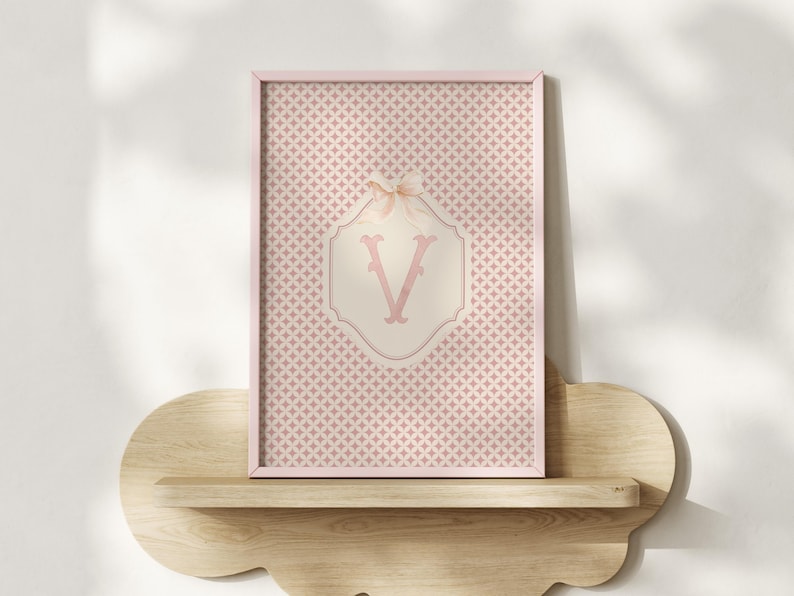 Puede incluir: Una impresi&oacute;n enmarcada con un fondo estampado rosa y blanco. La impresi&oacute;n tiene un marco rosa y un dise&ntilde;o central con la letra "V" en una fuente decorativa. Un peque&ntilde;o lazo adorna la parte superior del dise&ntilde;o. La impresi&oacute;n se muestra en una estanter&iacute;a de madera.