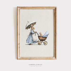 Puede incluir: Ilustración en acuarela de un conejo con un vestido azul y un paraguas, empujando un cochecito vintage con un conejito bebé. La obra de arte está enmarcada en un marco de madera marrón claro.