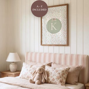Op de afbeelding: Een slaapkamer met een bed met een gestreepte hoofdbord en bloemenkussens. Een ingelijste print met de letter "K" en de tekst "A-Z INCLUDED" hangt aan de muur. Een pluchen konijn zit op het bed, wat de zachte esthetiek van de kamer aanvult.