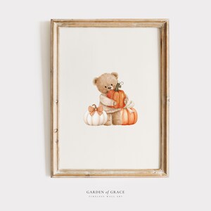 May include: A framed print featuring a watercolor illustration of a teddy bear hugging two pumpkins. The pumpkins are orange and white with a bow. The frame is a light brown color. The text "GARDEN OF GRACE TIMELESS WALL ART" is at the bottom.