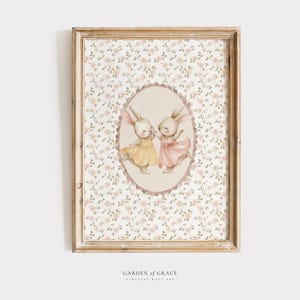 Op de afbeelding: Een ingelijste wandkunstprint met twee aquarelkonijntjes in jurken, tegen een bloemenachtergrond. De konijntjes staan in een centraal ovaal, één in een gele jurk en de ander in een roze jurk. De lijst is lichtbruin.