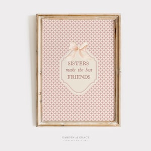 Może przedstawiać: Oprawiona grafika ścienna z różowym geometrycznym tłem. Grafika zawiera kremową etykietę z napisem "Sisters make the best FRIENDS" i kokardą. Rama jest w kolorze jasnobrązowym.
