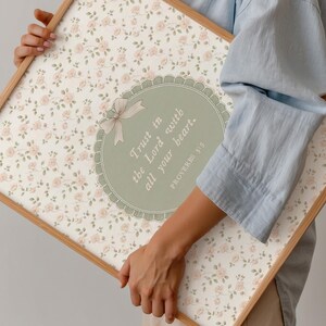 Op de afbeelding: Een ingelijste bloemenprint met een cr&egrave;mekleurige achtergrond en kleine roze rozen. De print heeft een centraal ovaal ontwerp met de tekst "Trust in the Lord with all your heart" en een strik. Het frame is gemaakt van licht hout.