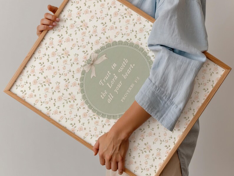 Op de afbeelding: Een ingelijste bloemenprint met een cr&egrave;mekleurige achtergrond en kleine roze rozen. De print heeft een centraal ovaal ontwerp met de tekst "Trust in the Lord with all your heart" en een strik. Het frame is gemaakt van licht hout.
