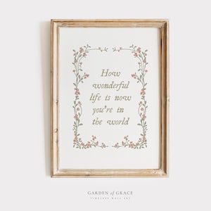 May include: A framed print with a light wood frame. The artwork displays the text "How wonderful life is now you're in the world" in a soft green font, framed by a floral border with pink flowers and green leaves. The words "Garden of Grace" are at the bottom.