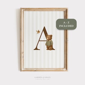 May include: Framed wall art featuring a brown letter "A" with a teddy bear wearing a green jacket. A small bird is flying near the letter. The background has vertical stripes in light beige and white. The text "A-Z INCLUDED" is in a green rounded rectangle.