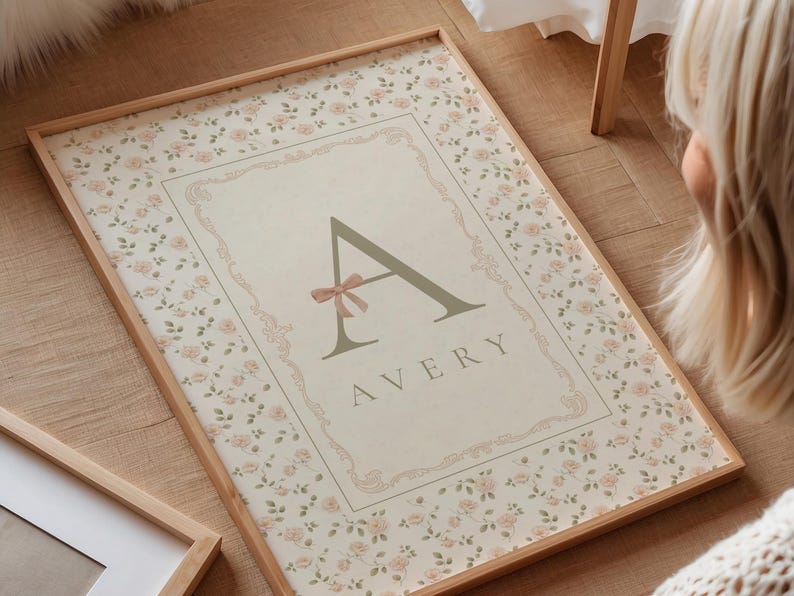 Pu&ograve; includere: Una stampa incorniciata con un bordo floreale e un design centrale con il nome "AVERY" sotto una grande lettera "A" ornata da un fiocco rosa. La cornice &egrave; in legno color chiaro.