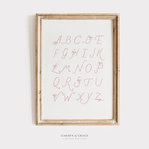May include: A framed print displaying the alphabet in a cursive style. The letters are a soft pink, each decorated with small bow details. The print is set against a cream background and framed in a light-toned, rustic wooden frame. The text "GARDEN OF GRACE" is at the bottom.