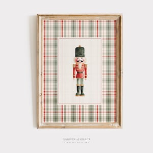 Könnte beinhalten: Gerahmter Druck mit einer Aquarellillustration eines Spielzeug-Nussknackersoldaten. Der Nussknacker trägt einen roten Mantel, schwarze Stiefel und einen schwarzen Hut. Der Hintergrund ist ein rot-grün-cremefarbenes Karomuster.