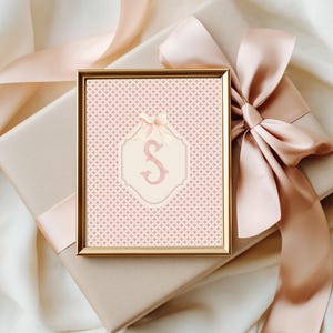 Puede incluir: Una impresi&oacute;n enmarcada con un borde dorado que presenta un fondo rosa estampado y un emblema color crema con la letra "S". La impresi&oacute;n est&aacute; encima de una caja de regalo envuelta con un gran lazo y cinta de color melocot&oacute;n.