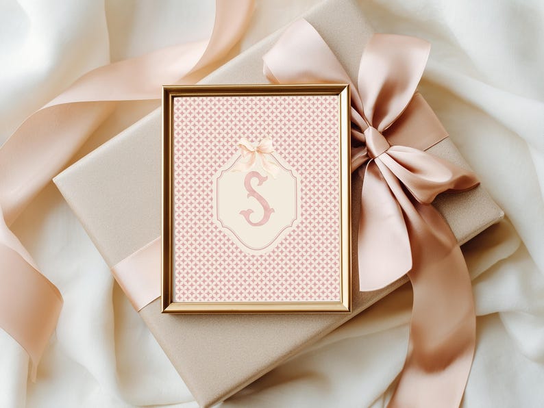 Puede incluir: Una impresi&oacute;n enmarcada con un borde dorado que presenta un fondo rosa estampado y un emblema color crema con la letra "S". La impresi&oacute;n est&aacute; encima de una caja de regalo envuelta con un gran lazo y cinta de color melocot&oacute;n.