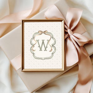 May include: A framed print featuring the letter "W" in a decorative, ornate frame. The artwork is in a gold frame and rests on a wrapped gift box with a large, peach-colored ribbon. The background is a soft, neutral color.