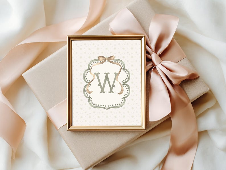May include: A framed print featuring the letter "W" in a decorative, ornate frame. The artwork is in a gold frame and rests on a wrapped gift box with a large, peach-colored ribbon. The background is a soft, neutral color.