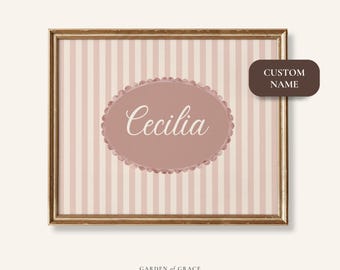 May include: A framed wall art print featuring a pink and white striped background. The print has a pink oval with the name "Cecilia" in white script. A brown label reads "CUSTOM NAME". The frame is gold.