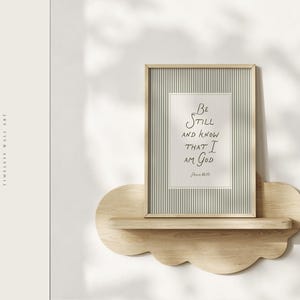 May include: A framed print with the text "Be Still and Know That I Am God" on a striped background, displayed on a cloud-shaped wooden shelf. The frame is light-colored, and the shelf is made of natural wood. The words "Garden of Grace" are on the left.