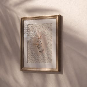 K&ouml;nnte beinhalten: Gerahmtes Kunstwerk mit einer detaillierten Illustration eines Hasen, der Blumen h&auml;lt. Der Hase befindet sich vor einem floralen Hintergrund in einer ovalen Form. Der Rahmen ist hellbraun, und das Kunstwerk h&auml;ngt an einer beigefarbenen Wand.
