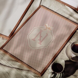 Puede incluir: Una impresi&oacute;n enmarcada con un dise&ntilde;o de patr&oacute;n rosa y un emblema blanco y rosa claro. El emblema muestra la letra "N" en una fuente decorativa. Una taza de t&eacute; y una crema se colocan cerca del marco.