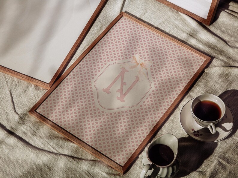 Puede incluir: Una impresi&oacute;n enmarcada con un dise&ntilde;o de patr&oacute;n rosa y un emblema blanco y rosa claro. El emblema muestra la letra "N" en una fuente decorativa. Una taza de t&eacute; y una crema se colocan cerca del marco.
