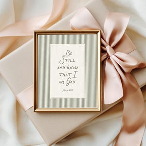 May include: A framed print with the text "Be Still and Know That I Am God" in elegant script. The print has a gold frame and is set against a backdrop of a gift box with a peach-colored ribbon and bow.