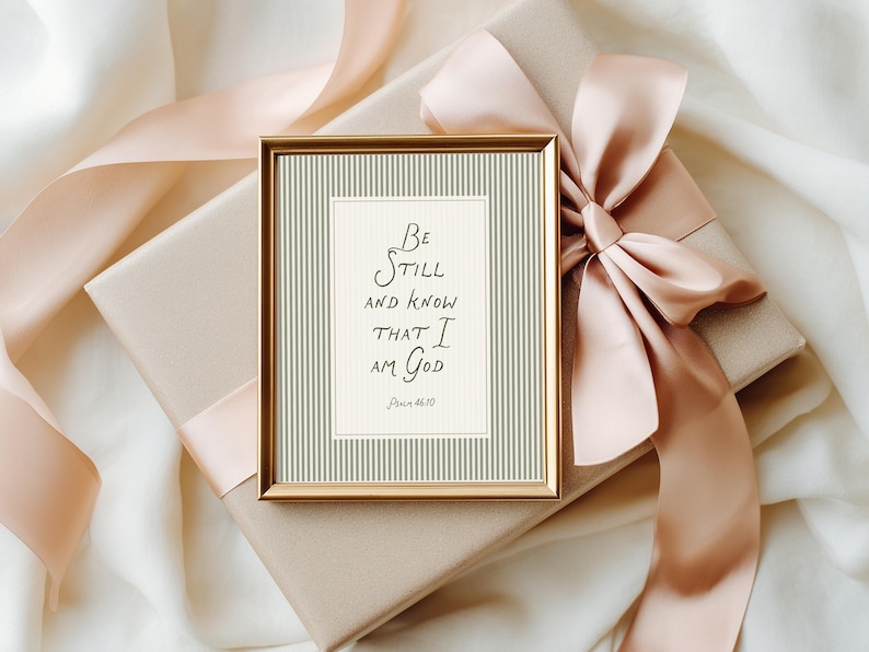 May include: A framed print with the text "Be Still and Know That I Am God" in elegant script. The print has a gold frame and is set against a backdrop of a gift box with a peach-colored ribbon and bow.