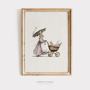Puede incluir: Impresión en acuarela enmarcada de un conejo con un vestido rosa y un paraguas, empujando un cochecito vintage con otro conejo dentro. La obra de arte está en un marco de madera marrón claro. El texto "GARDEN OF GRACE" y "TIMELESS WALL ART" está en la parte inferior.