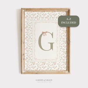 May include: A framed print with a floral background and a central letter "G" in green, decorated with a pink bow. The frame is light brown. The artwork includes the text "A-Z INCLUDED" and "GARDEN OF GRACE".