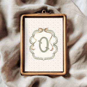 May include: A framed print featuring the letter "Q" in a soft green hue, set against a dotted background. The letter is framed by a decorative border with a bow at the top. The frame is gold-colored.