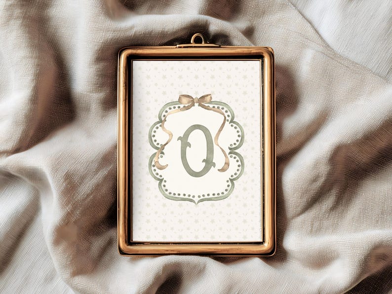 May include: A framed print featuring the letter "Q" in a soft green hue, set against a dotted background. The letter is framed by a decorative border with a bow at the top. The frame is gold-colored.
