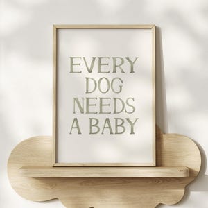 Op de afbeelding: Een ingelijste print met de tekst "EVERY DOG NEEDS A BABY" in een zachte groene tint. De print wordt weergegeven op een licht houten plank met een decoratieve, wolkvormige vorm.