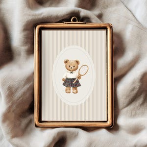 K&ouml;nnte beinhalten: Gerahmter Druck mit einer Aquarellillustration eines Teddyb&auml;ren in Tennisbekleidung, der einen Schl&auml;ger h&auml;lt. Der B&auml;r befindet sich vor einem gestreiften Hintergrund in einem ovalen Rahmen. Der Rahmen ist goldfarben.