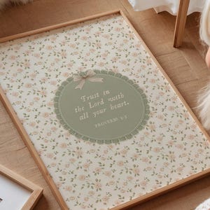 Op de afbeelding: Een ingelijste print met een bloemenpatroon achtergrond en de tekst "Trust in the Lord with all your heart." De print heeft een lichtbruine houten lijst en een decoratief ovaal met een strik.