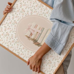 May include: A framed artwork featuring a floral pattern of small pink roses on a cream background. The artwork has a light brown wooden frame. A central design element includes a stack of books tied with a pink ribbon, set within a light peach circle.
