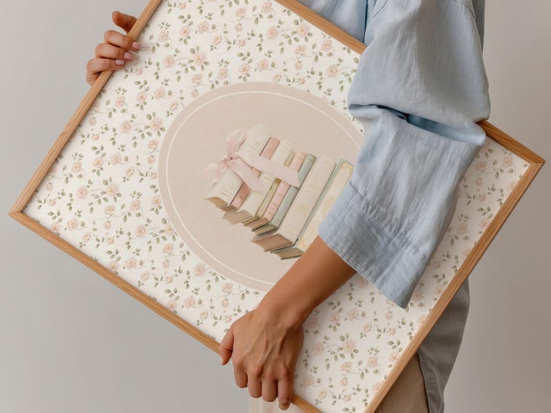 May include: A framed artwork featuring a floral pattern of small pink roses on a cream background. The artwork has a light brown wooden frame. A central design element includes a stack of books tied with a pink ribbon, set within a light peach circle.
