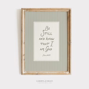 May include: Framed wall art with a light wood frame. The artwork features a cream-colored background with a vertical striped pattern in light green. The text "Be Still and Know That I Am God" is written in black script.