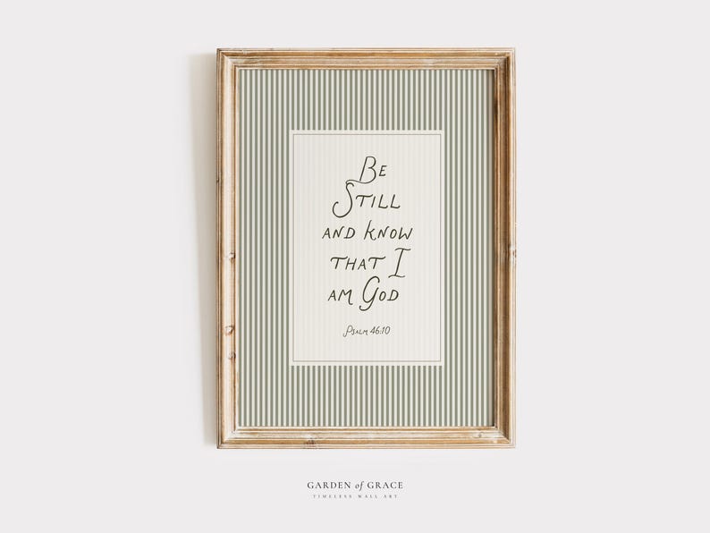 May include: Framed wall art with a light wood frame. The artwork features a cream-colored background with a vertical striped pattern in light green. The text "Be Still and Know That I Am God" is written in black script.