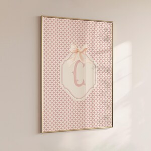 Puede incluir: Impresi&oacute;n enmarcada con un patr&oacute;n geom&eacute;trico rosa. Un lazo rosa claro adorna un emblema crema con la letra "C" en rosa a juego. El marco es dorado. La impresi&oacute;n est&aacute; colgada en la pared.