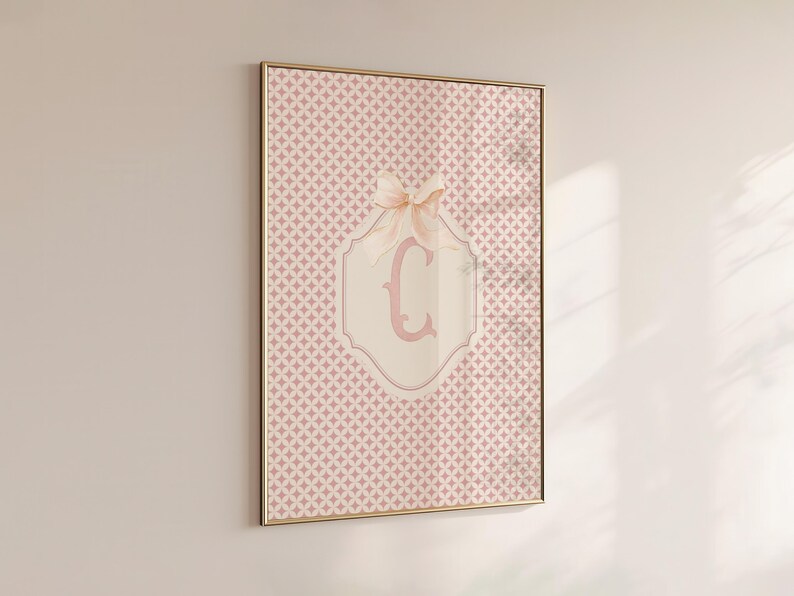 Puede incluir: Impresi&oacute;n enmarcada con un patr&oacute;n geom&eacute;trico rosa. Un lazo rosa claro adorna un emblema crema con la letra "C" en rosa a juego. El marco es dorado. La impresi&oacute;n est&aacute; colgada en la pared.