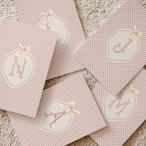 Puede incluir: Tarjetas rosas estampadas con un dise&ntilde;o geom&eacute;trico repetitivo. Cada tarjeta presenta un emblema color crema con una letra rosa y un lazo. Las letras visibles son A, J, M y N. Las tarjetas est&aacute;n dispuestas sobre una superficie de color neutro.