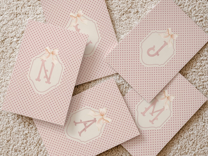 Puede incluir: Tarjetas rosas estampadas con un dise&ntilde;o geom&eacute;trico repetitivo. Cada tarjeta presenta un emblema color crema con una letra rosa y un lazo. Las letras visibles son A, J, M y N. Las tarjetas est&aacute;n dispuestas sobre una superficie de color neutro.